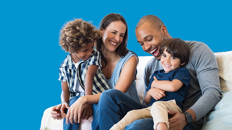 Mulit-racial family of four sits on couch laughing and enjoying each other's company with a cutout bright blue background.